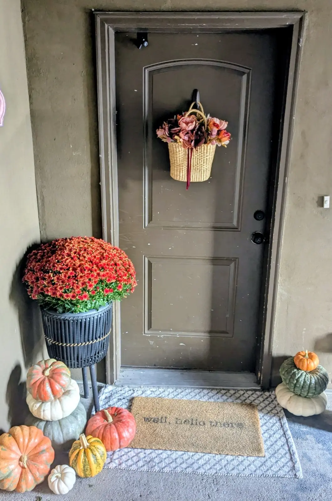 Apartment doorway with a basket door hanger, a mum on a black planter stand, and stacked pumpkins